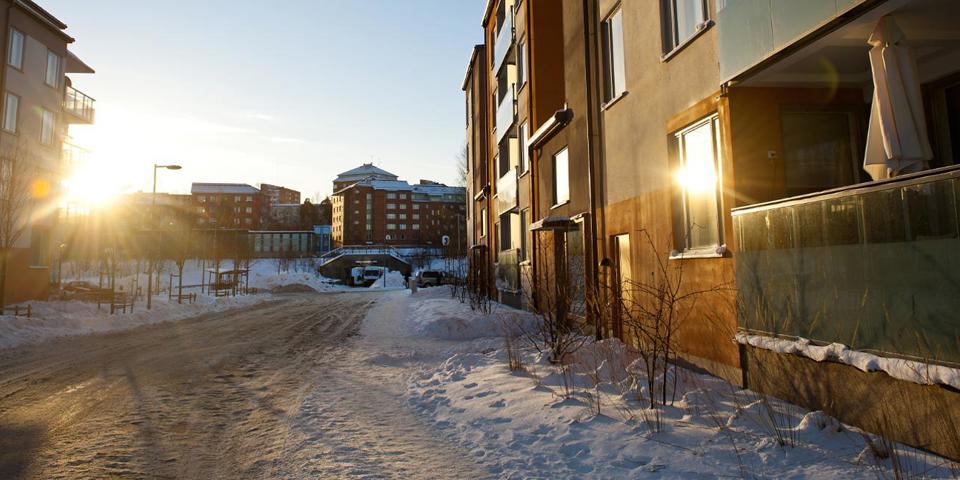 En gata med flerbostadshus på vardera sidan och med en pendeltågsstation i bakgrunden. Det är vinter och gatan är snöröjd. Solen ligger lågt och skiner mellan husen. Foto.
